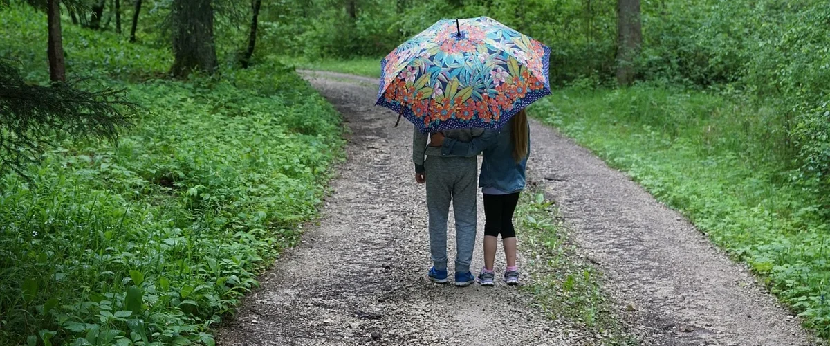 Deux enfants vus de dos sous un parapluie à fleurs sur un chemin, que faire quand il pleut avec les enfants